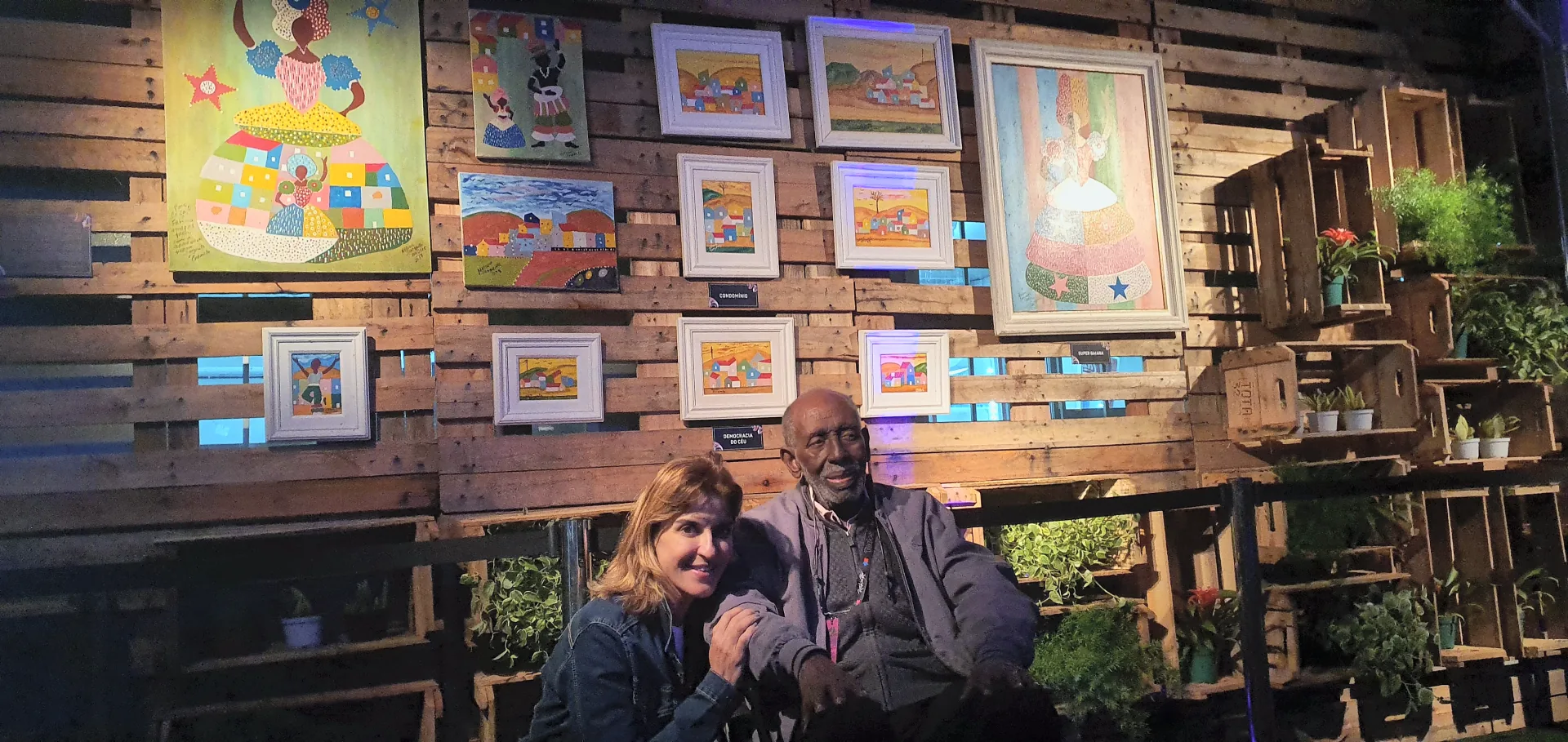 Nelson Sargento diante de pinturas expostas, acompanhado por uma mulher.